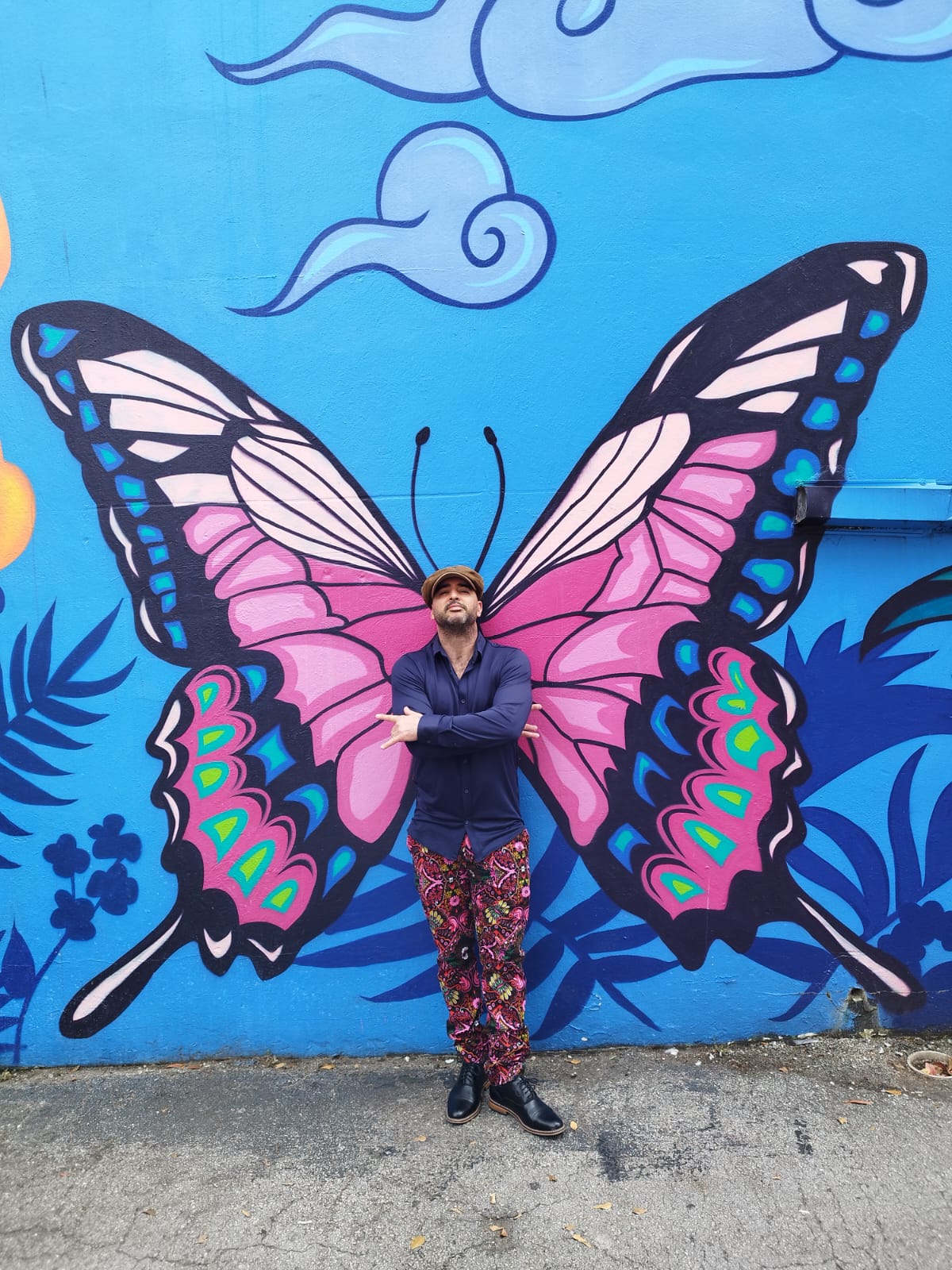Person standing in front of a colorful butterfly mural with arms crossed, wearing a blue jacket and patterned pants.