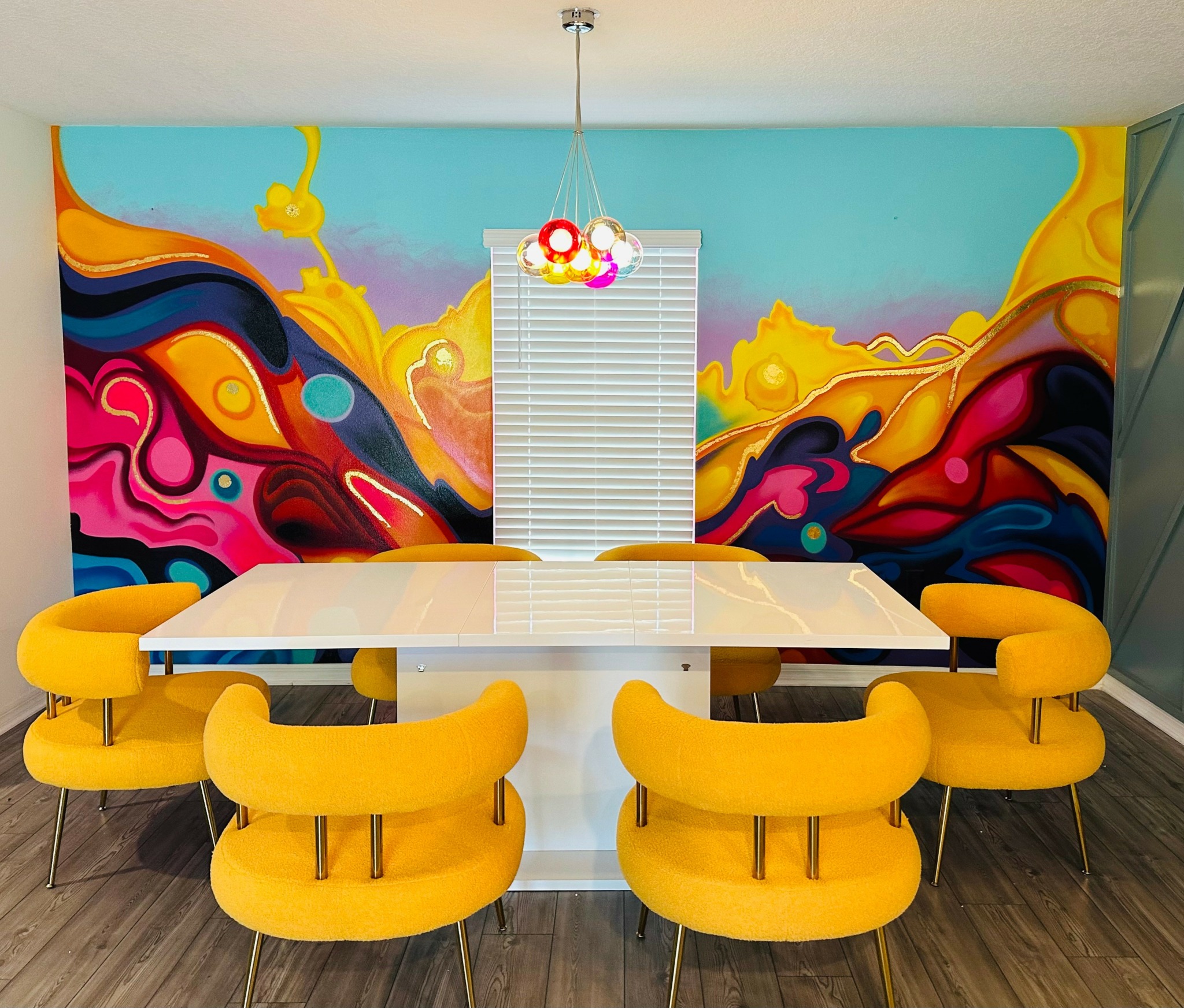 Colorful abstract mural behind a white table with six yellow chairs, a window with blinds, and a hanging light fixture.