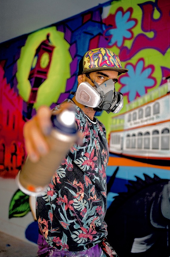 Person wearing a colorful cap, mask, and floral shirt pointing a spray can at the camera, graffiti background.