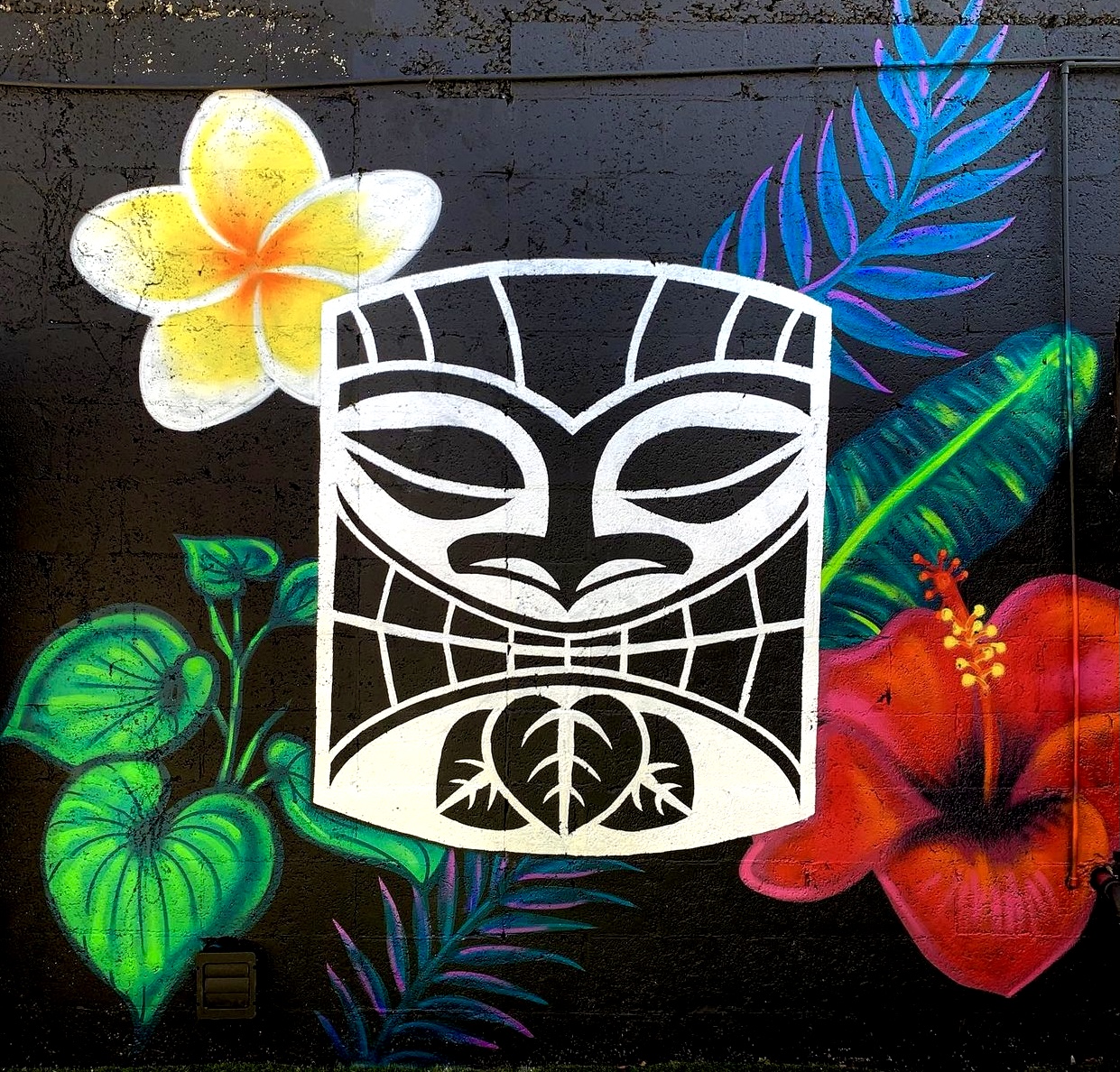Colorful mural with a white tiki face in front of tropical flowers and leaves on a black background.