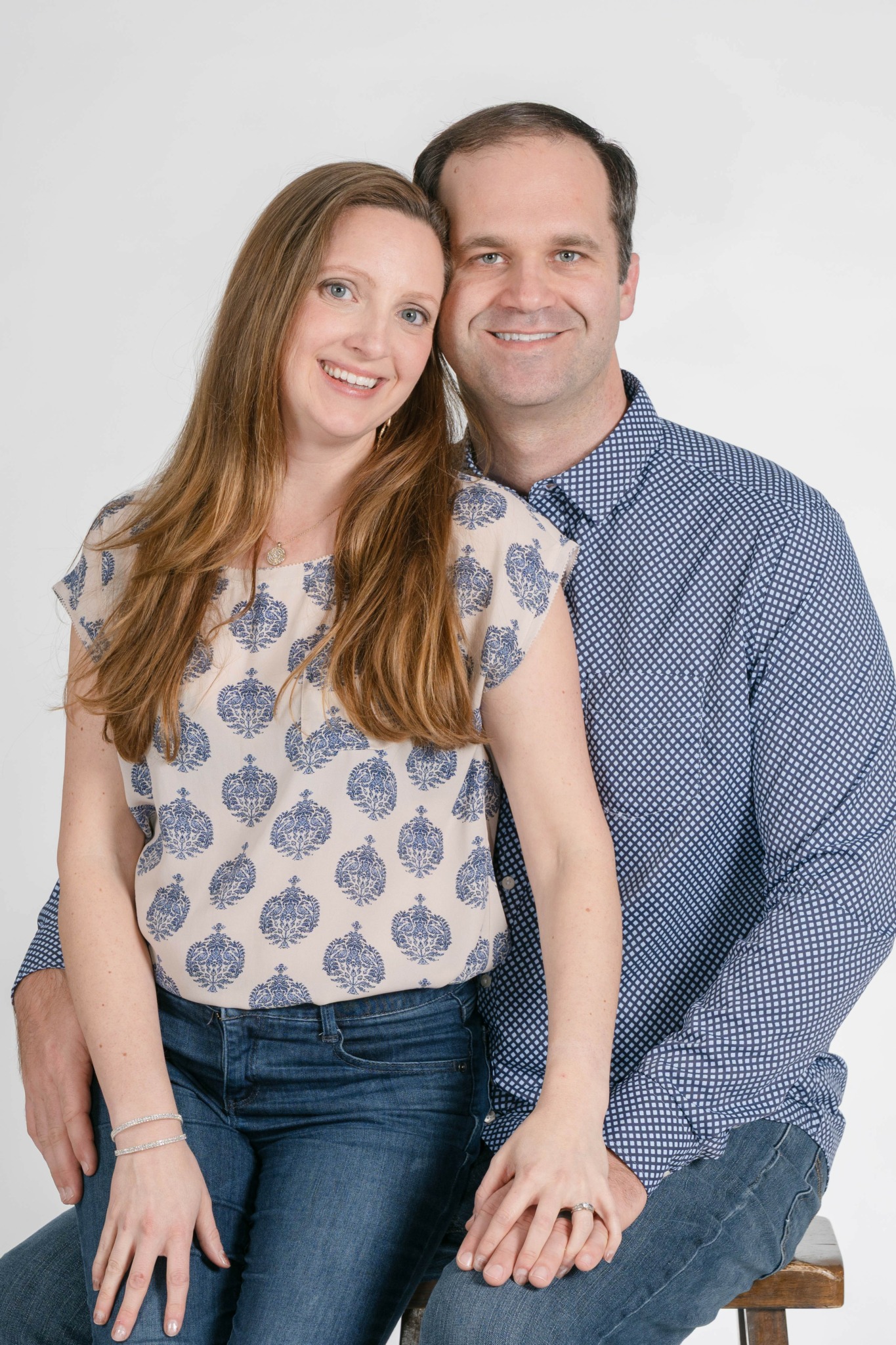 A smiling woman with long hair and a man sit close together, both looking at the camera against a plain background.