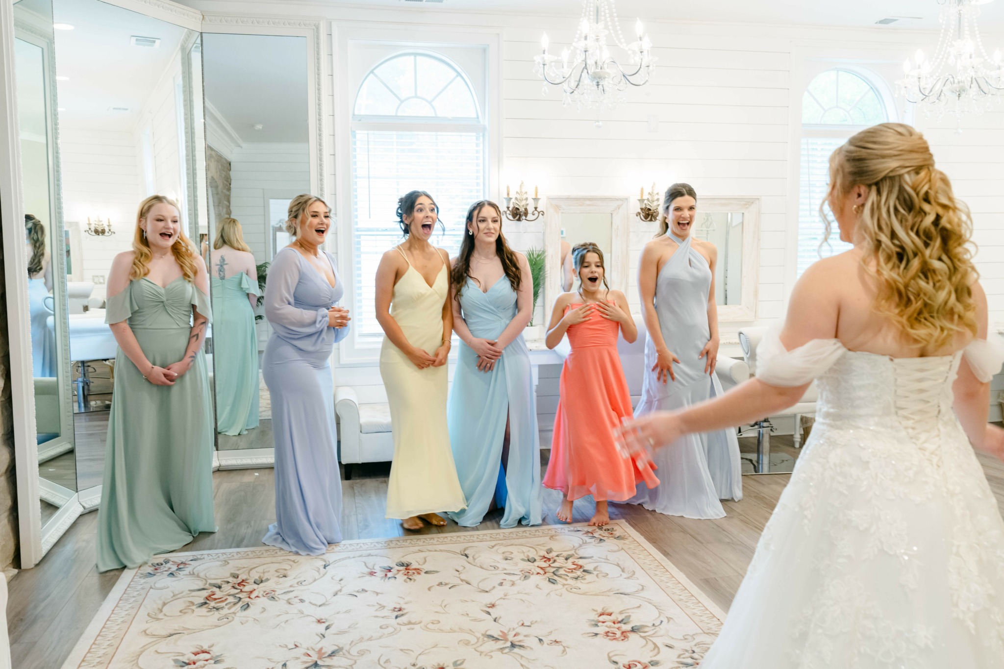 Bride in white dress facing seven women in pastel dresses in a bright room with large windows and chandelier.