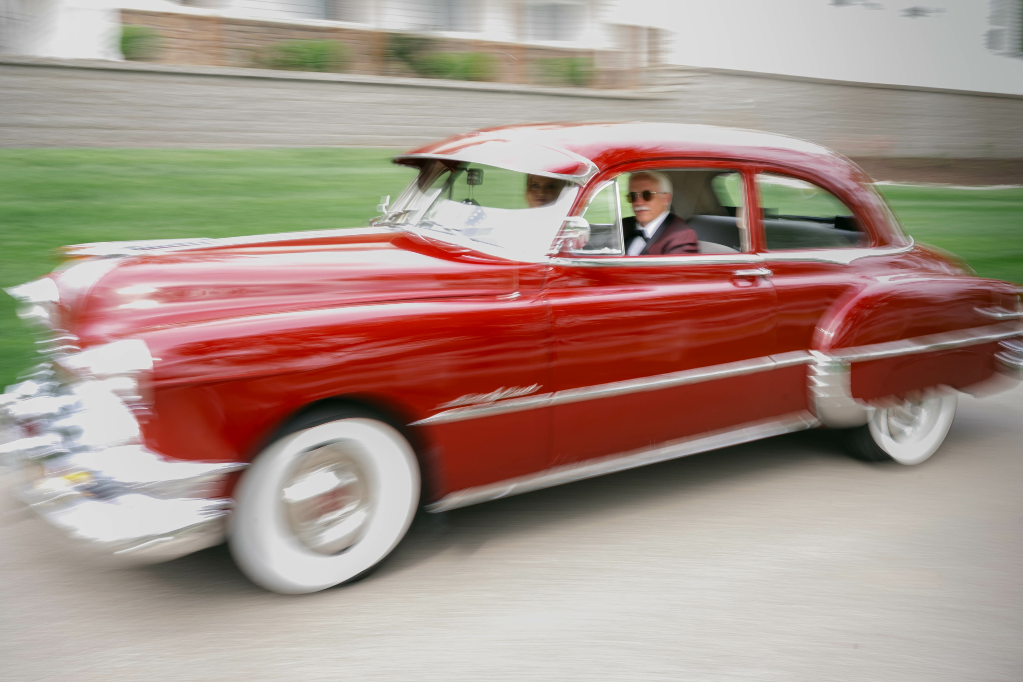 A person in sunglasses driving a vintage red car on a street, with motion blur indicating speed.
