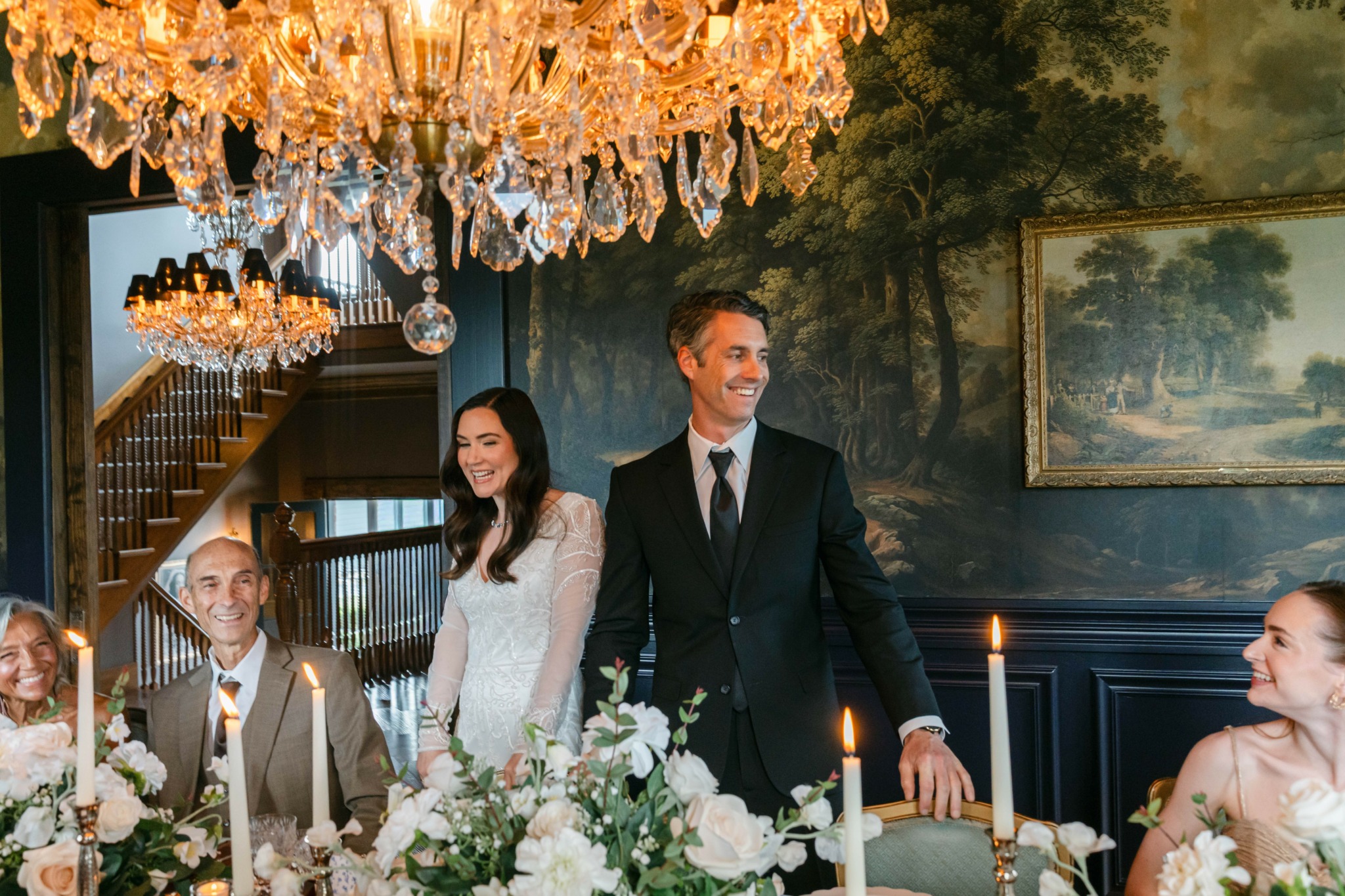 People in formal attire gather around a decorated table with candles and flowers, under a chandelier in a decorated room.