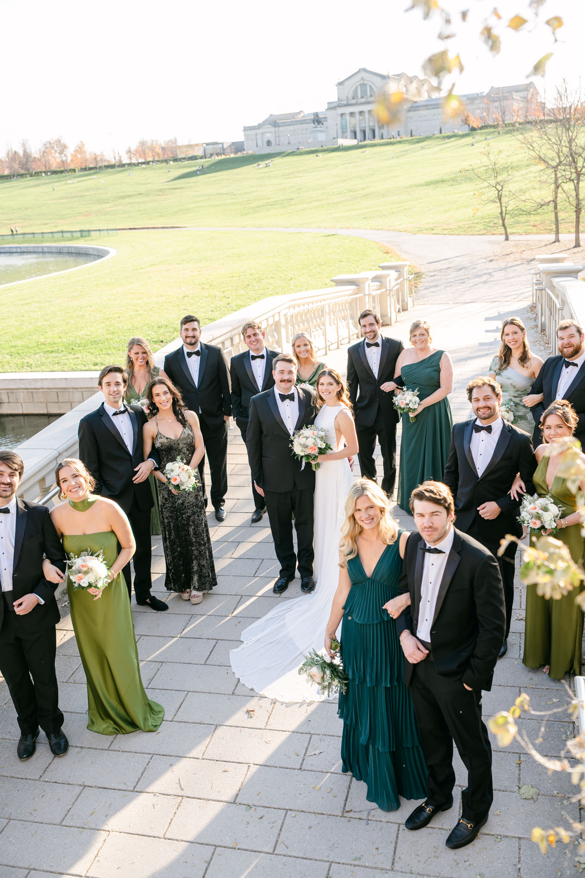 Group of people dressed in formal attire outdoors on a paved area with a grassy landscape and buildings in background.