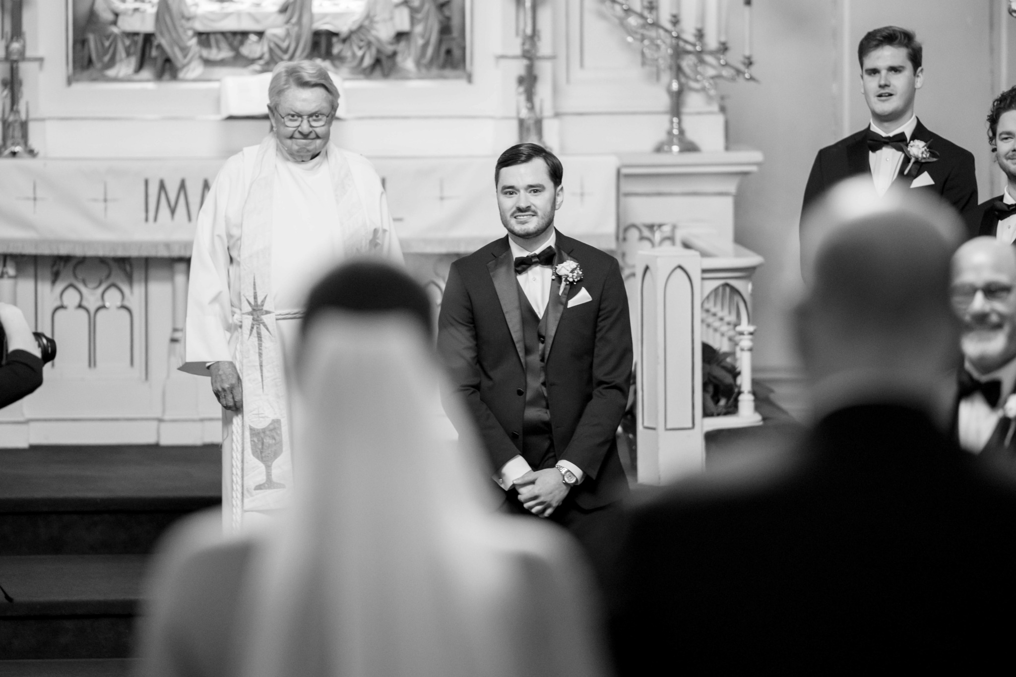 A wedding ceremony with a groom, officiant, and guests in a church setting, black and white photo, focus on groom.