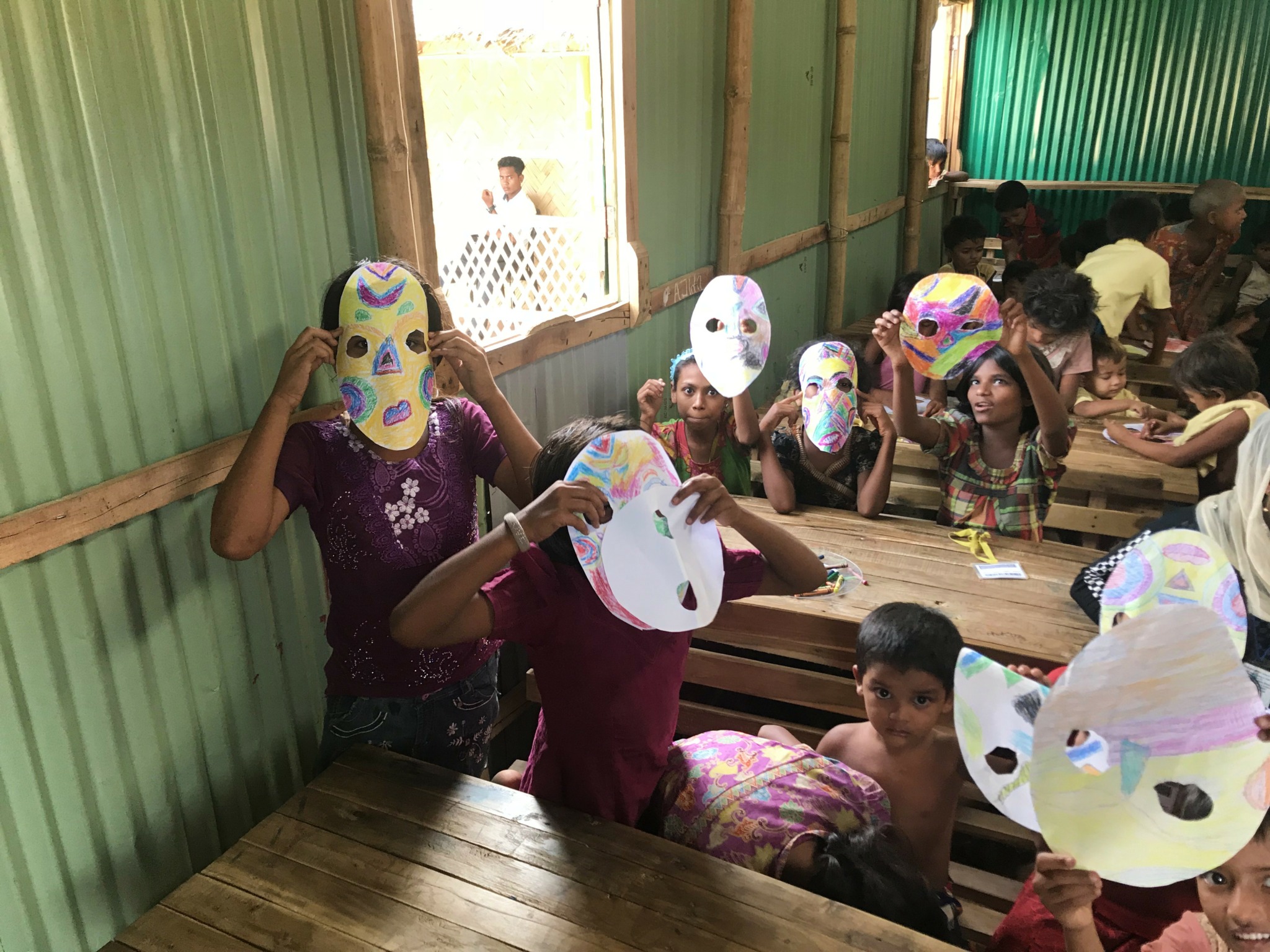 Children inside a green-walled room wearing paper masks, sitting and standing around wooden tables, some looking at the camera.