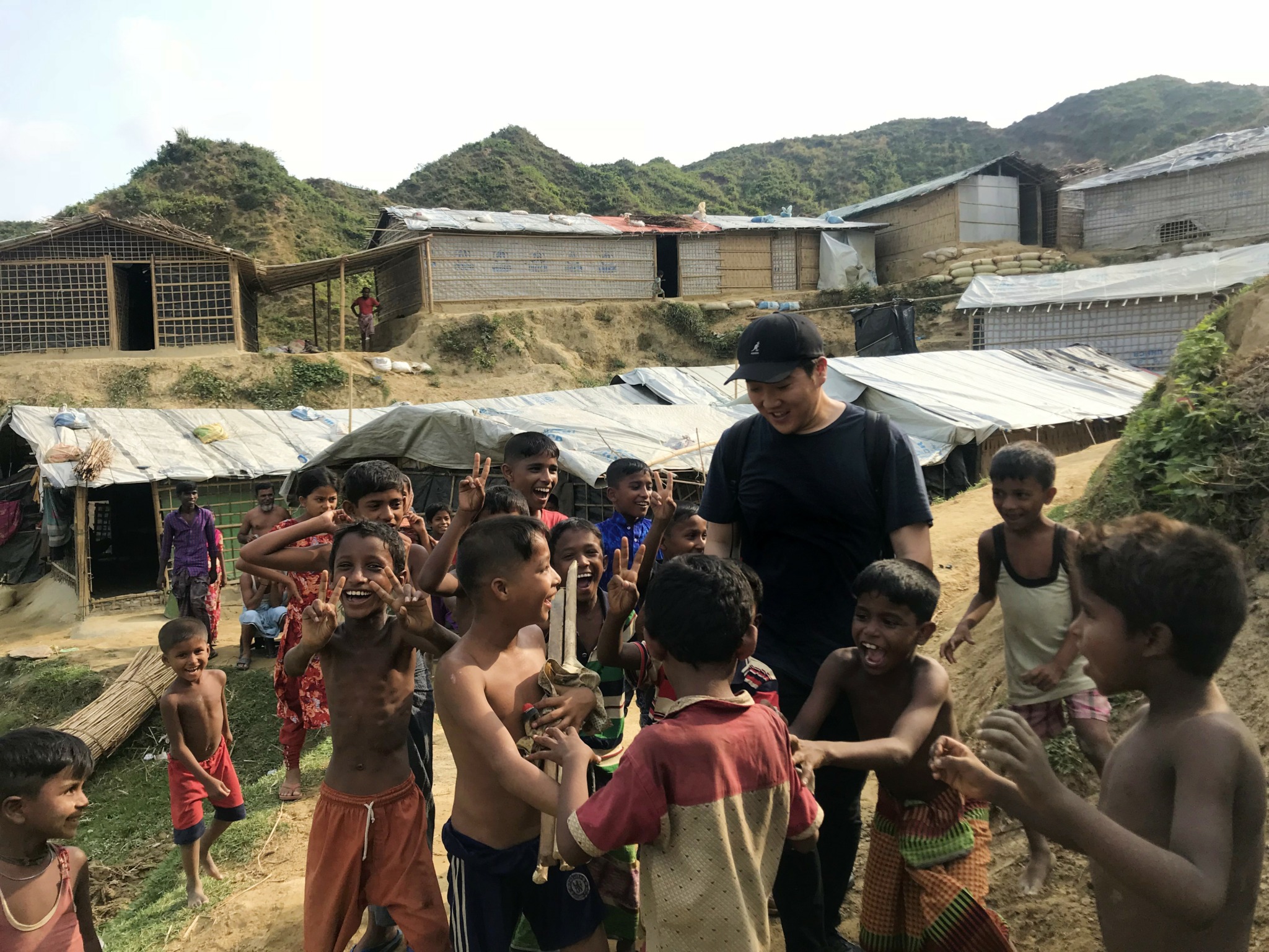 Group of children and a person outdoors in a rural area with hills and buildings in the background.