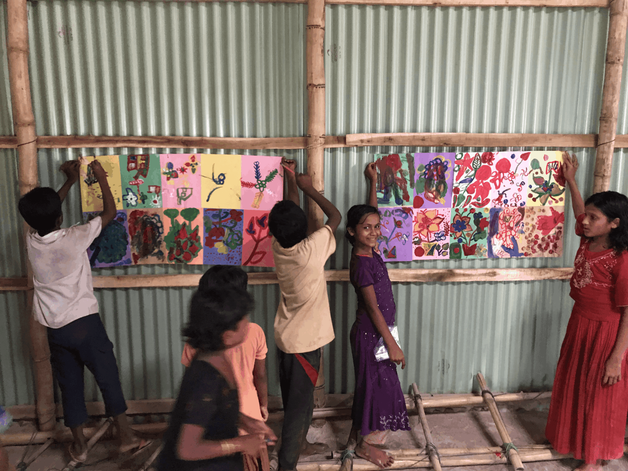 Four children stand in front of colorful artwork displayed on a corrugated metal wall, some reaching to adjust or view the art.
