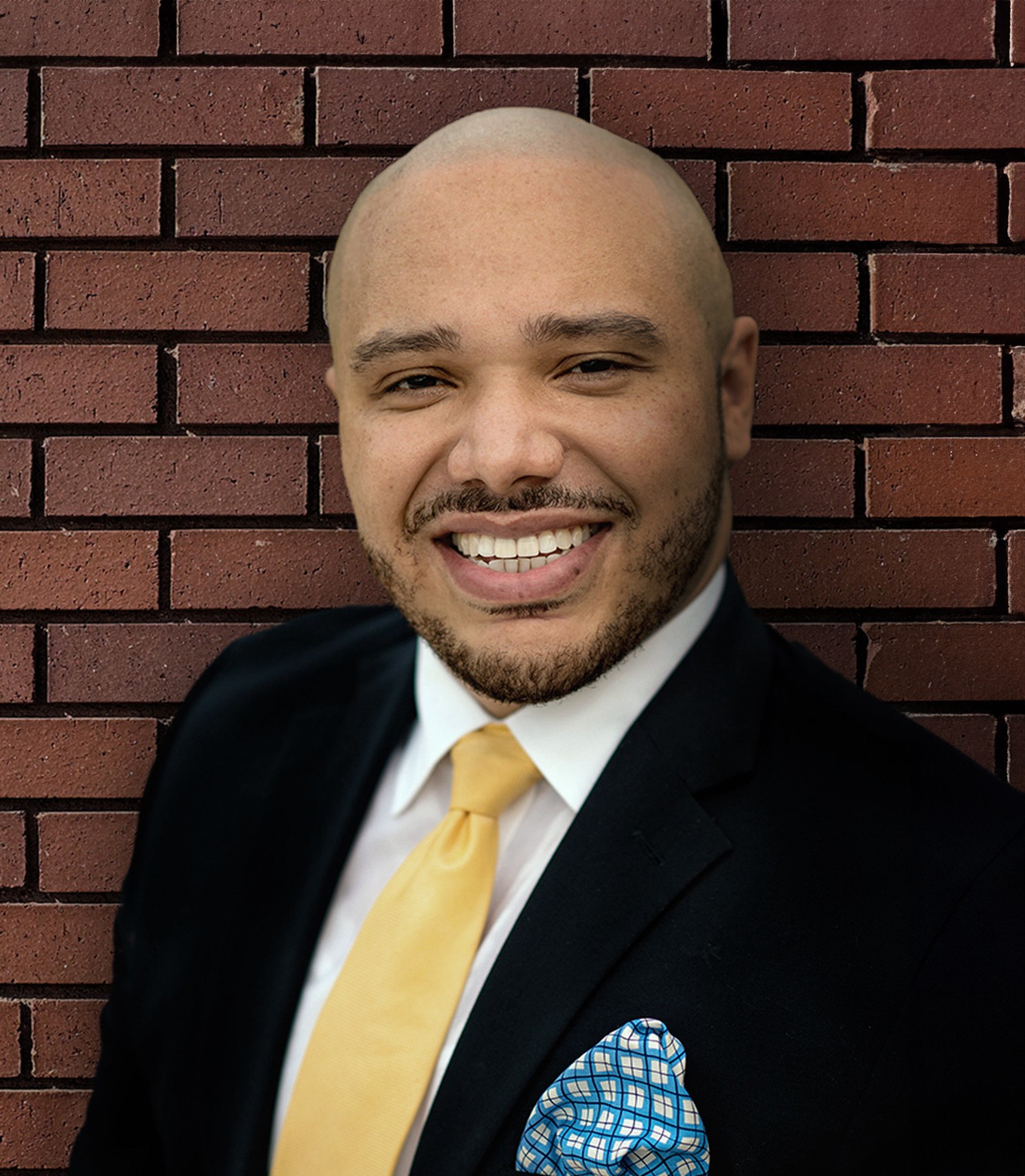 Man with a bald head, smiling, wearing a black suit, white shirt, yellow tie, and a patterned pocket square, standing in front of a brick wall.