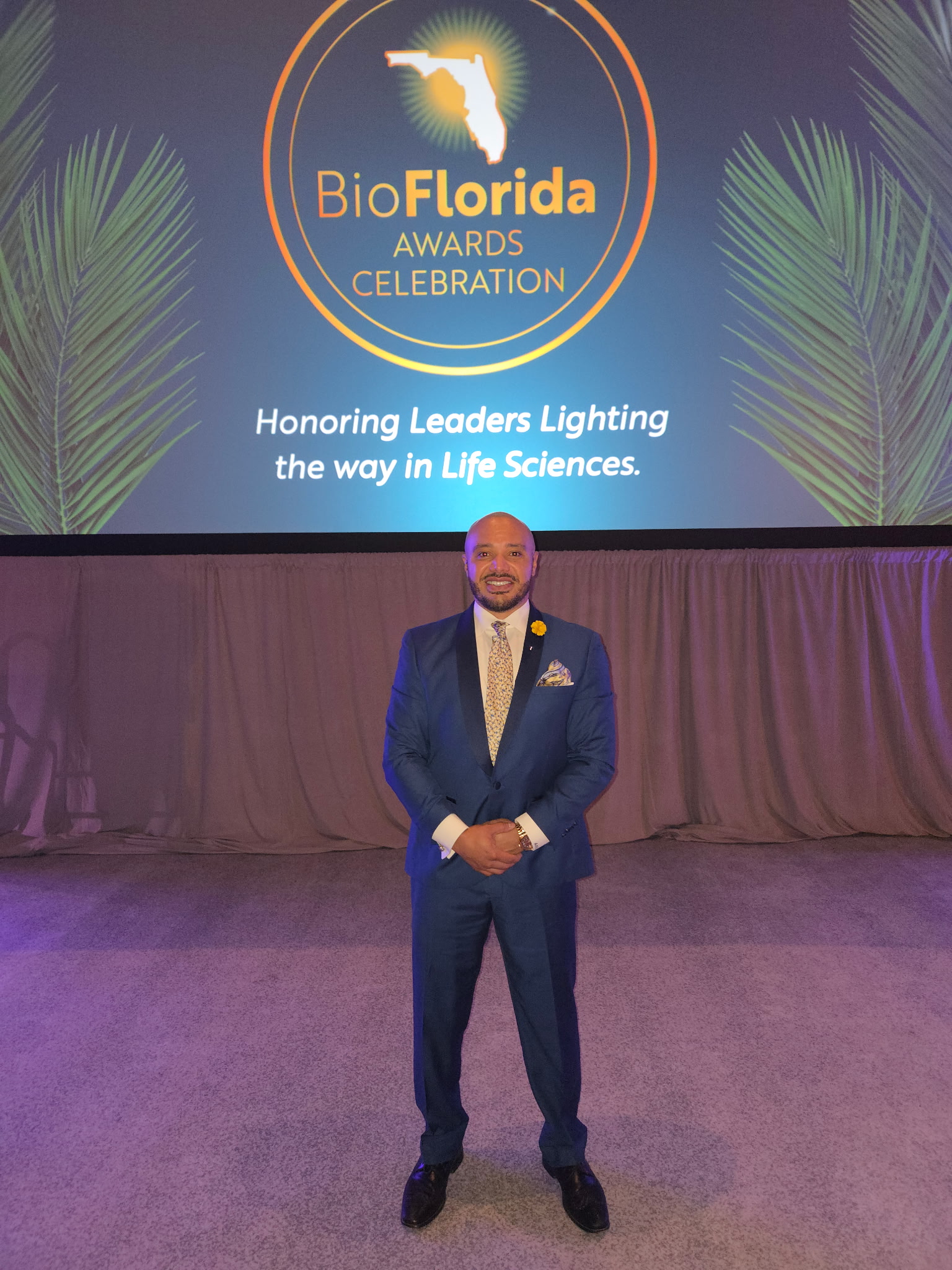 Man in a suit standing on stage with a large screen behind him displaying BioFlorida awards celebration logo.