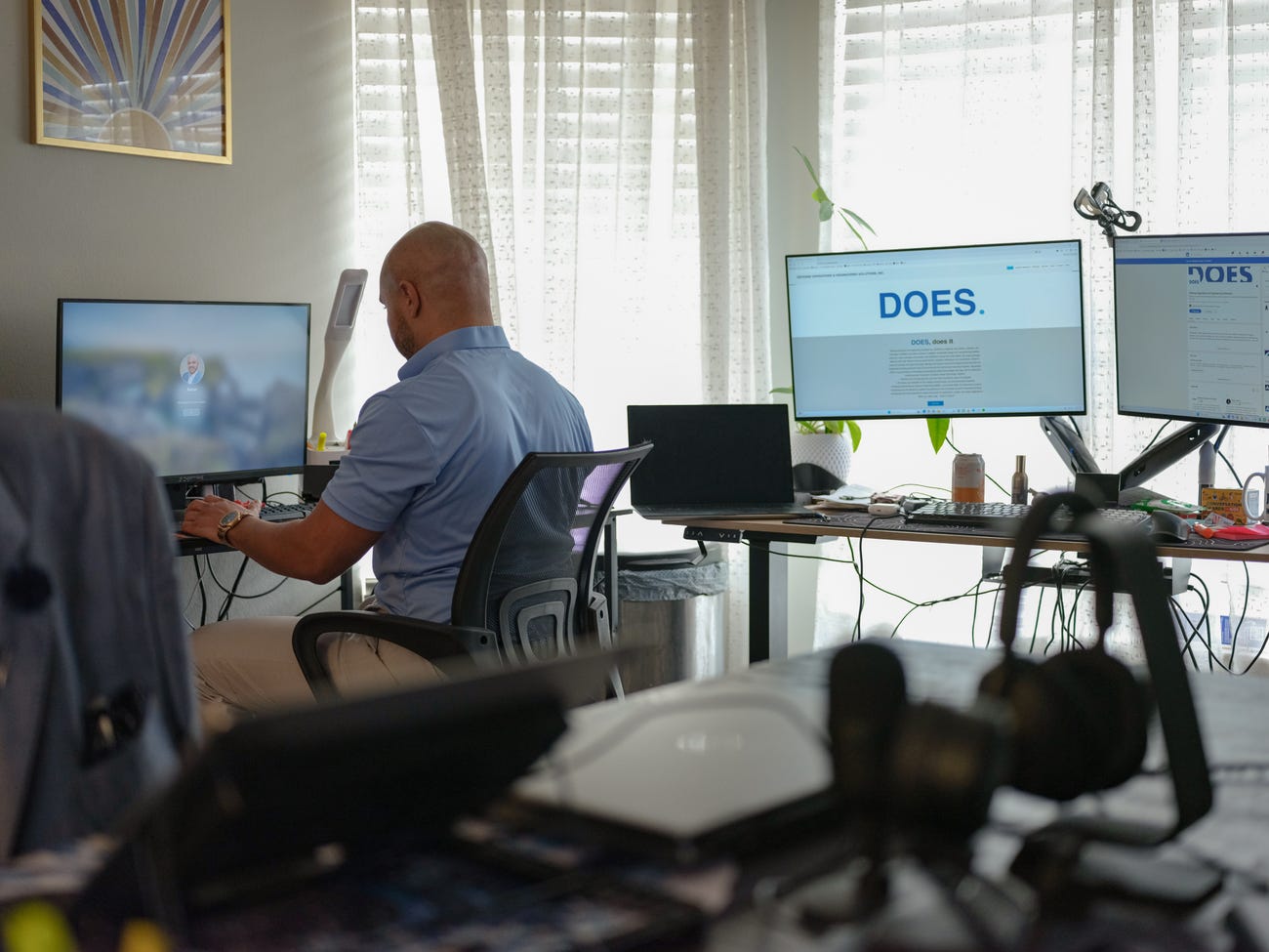 Bald man working at desk with multiple monitors in a bright office space.