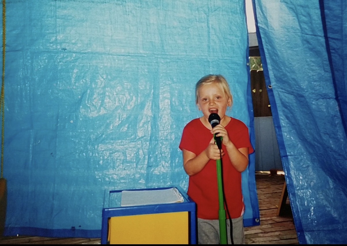 Child with blonde hair in red shirt holding a microphone on stage with blue curtains behind.