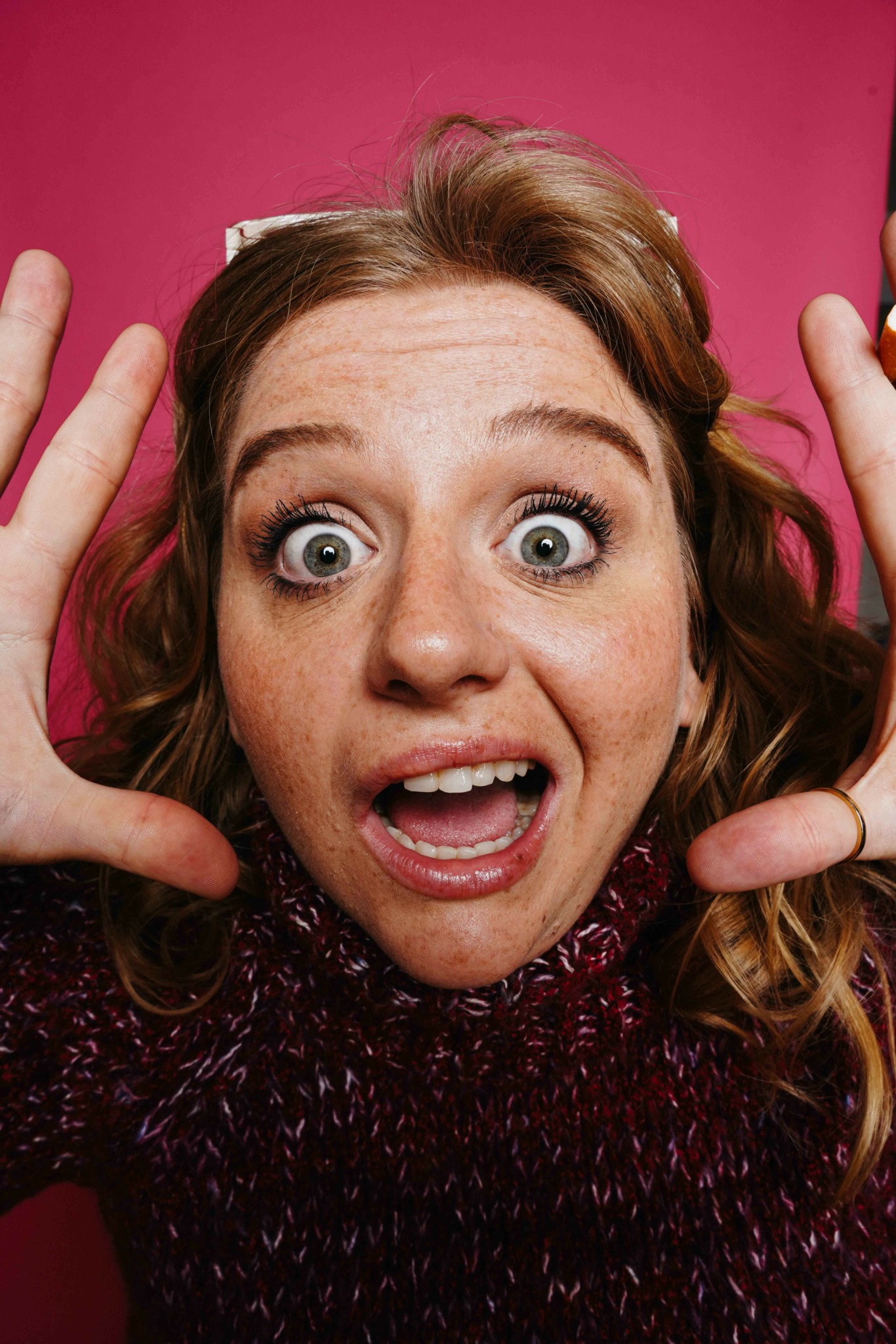 Woman with wide eyes and open mouth, hands near face, against pink background.