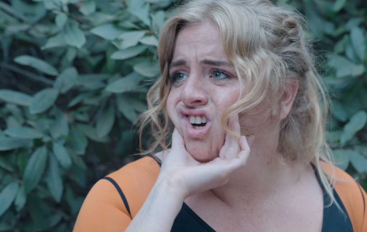 Woman with blonde curly hair making a distressed facial expression, holding her face with her hand, outdoors with green foliage.