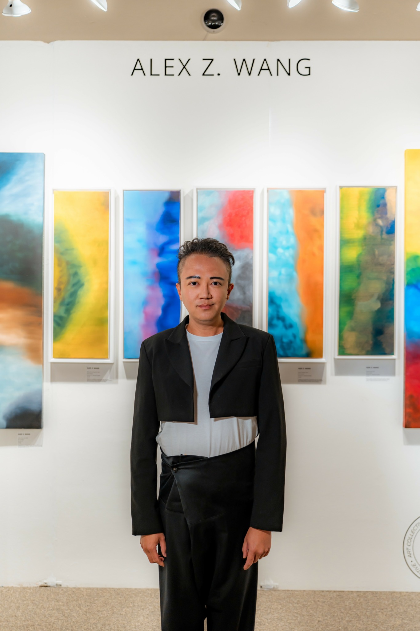 Young man in black suit and white shirt standing in front of colorful abstract artwork at an art gallery.