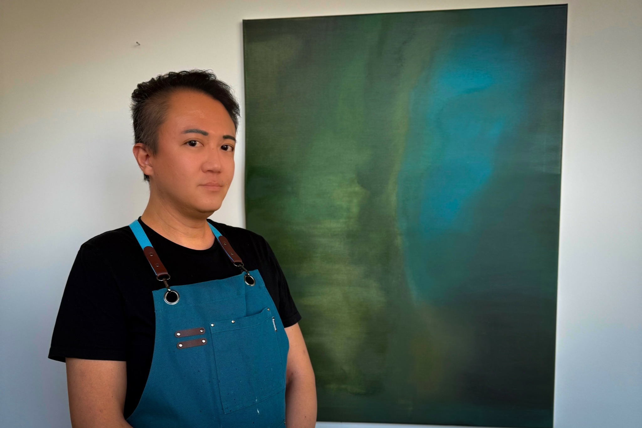 Person with short hair wearing a black shirt and blue apron standing next to an abstract green and blue painting.