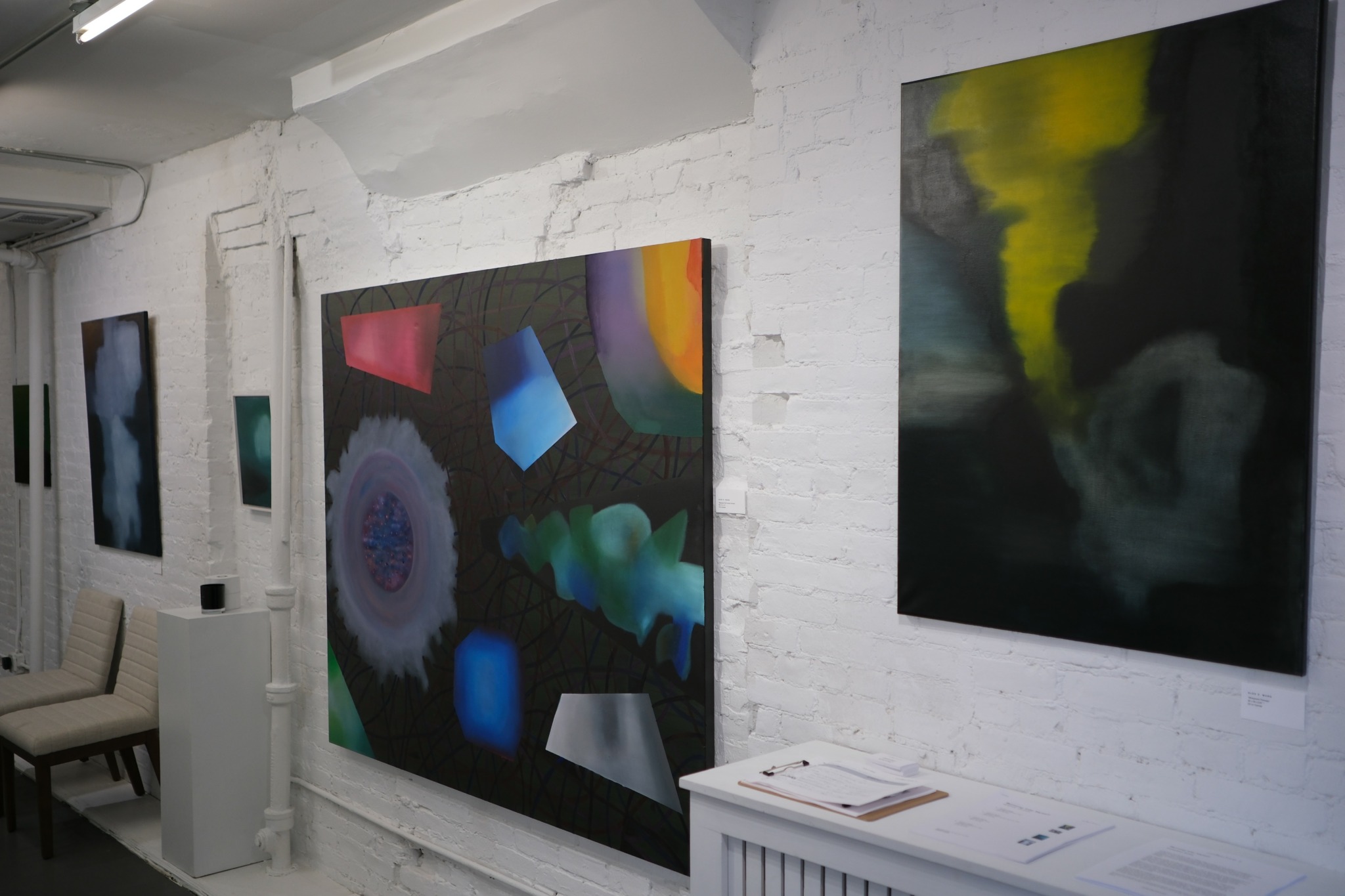 Gallery wall with abstract colorful paintings on white brick wall, seating area with chairs, and white furniture.