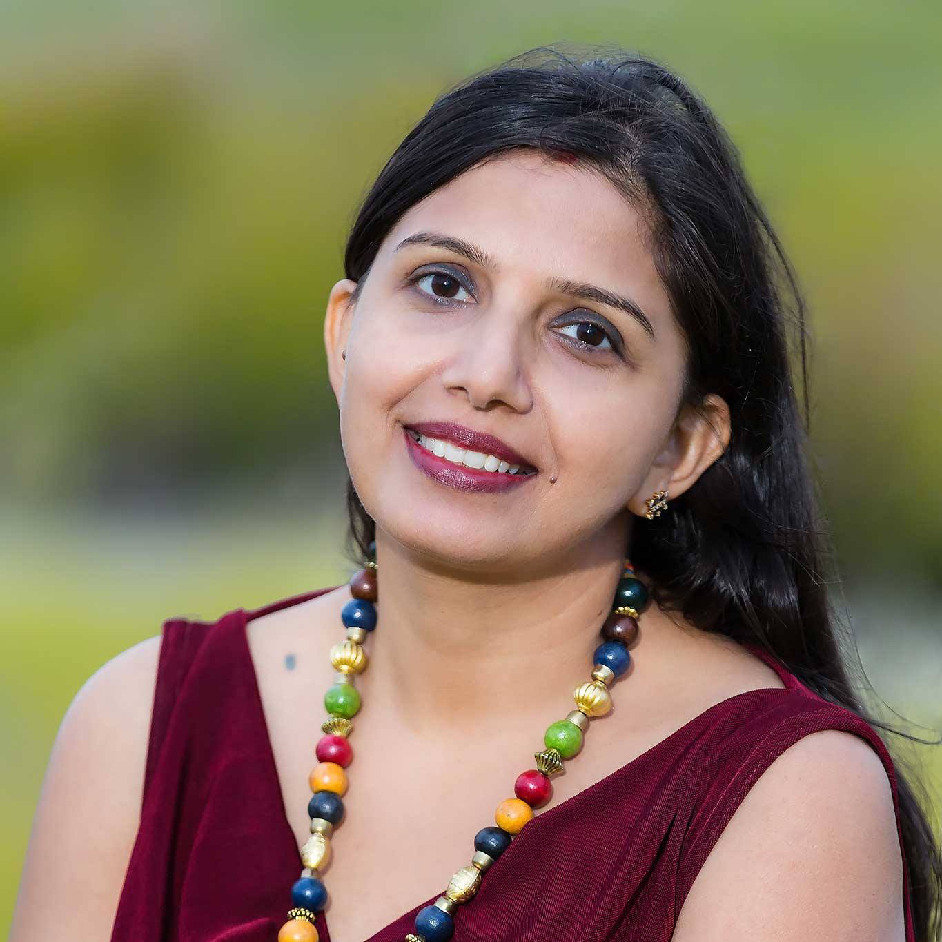 Woman with long dark hair smiling outdoors, wearing a maroon top and colorful beaded necklace.