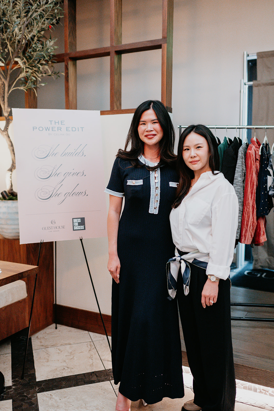 Two women standing indoors, smiling, with a clothing rack and a sign in the background.