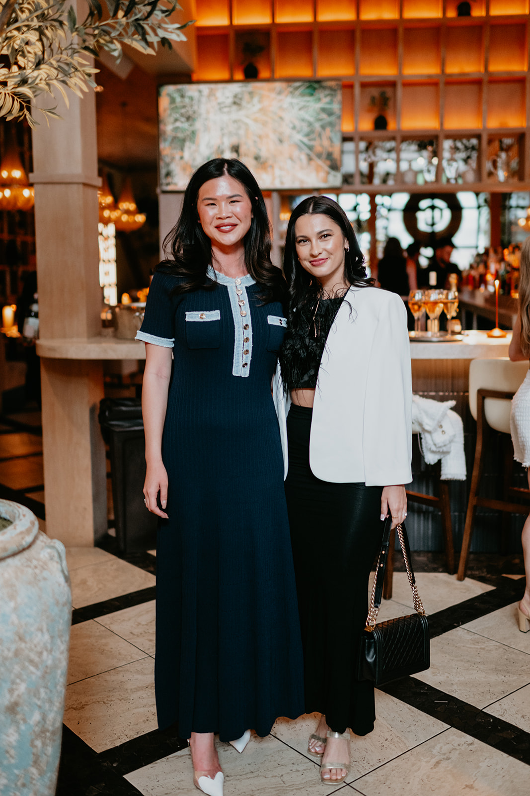 Two women standing together in a warmly lit indoor setting, smiling at the camera, with a bar and decorative elements in the background.