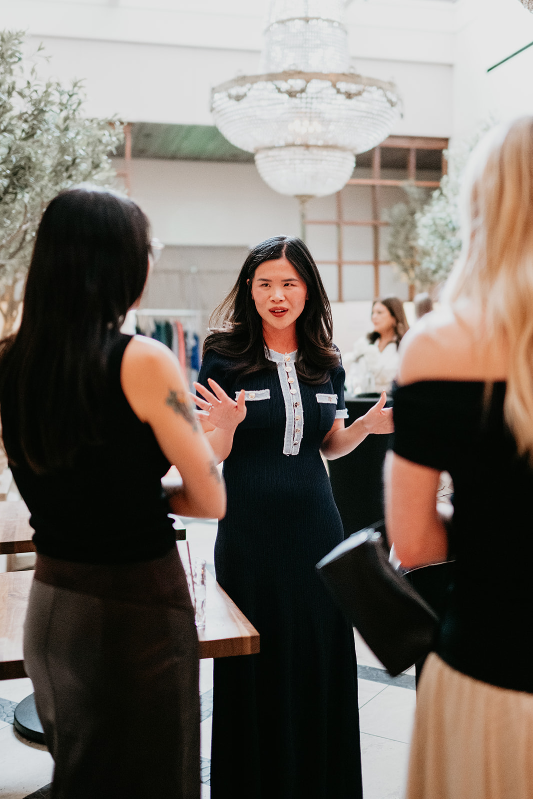 Three women engaged in conversation indoors with a chandelier and large windows in the background.