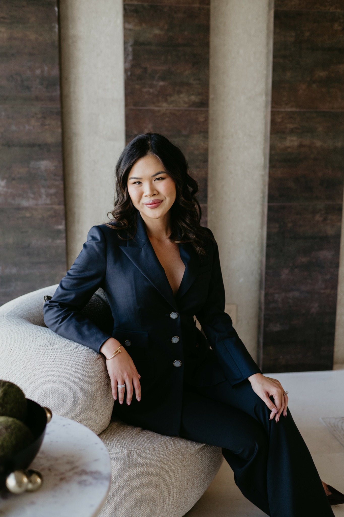 Woman in dark suit sitting on a light-colored sofa with wooden and concrete background.