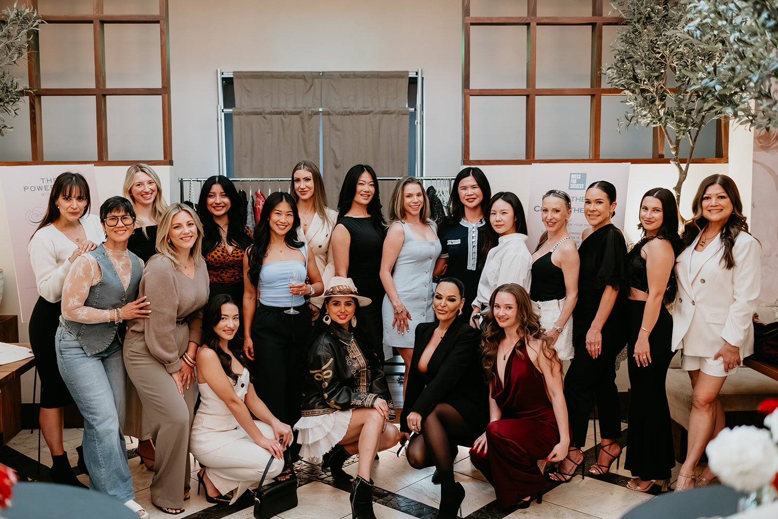 Group of 20 women and girls standing together indoors, smiling, in front of a wall with wooden panels and plants.
