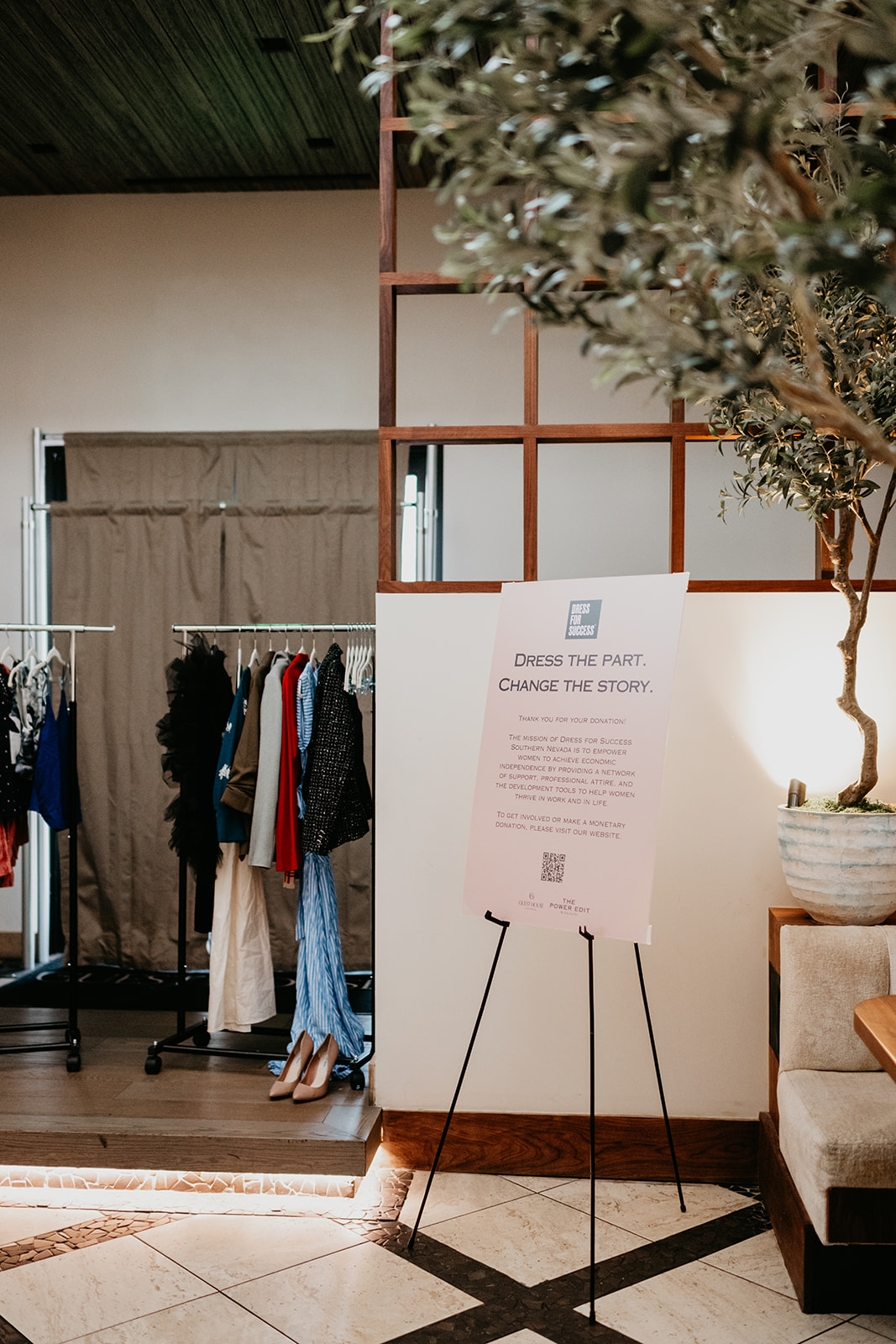 Clothing rack with various garments, a sign on an easel, and a potted tree in an indoor space.