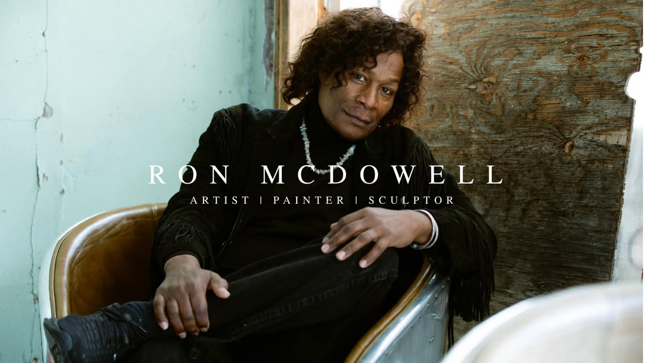 Portrait of Ron McDowell sitting in a chair, with a wooden wall background and a light blue wall behind.