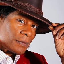 Close-up of a person wearing a brown hat and red jacket, holding the brim of the hat with their right hand.
