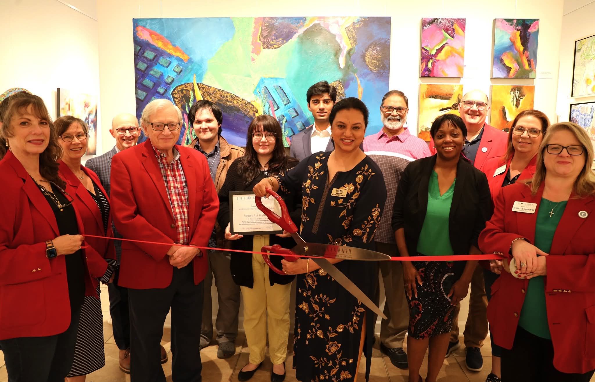 Group of people at an art gallery opening, holding a ribbon and certificate, with colorful paintings in the background.