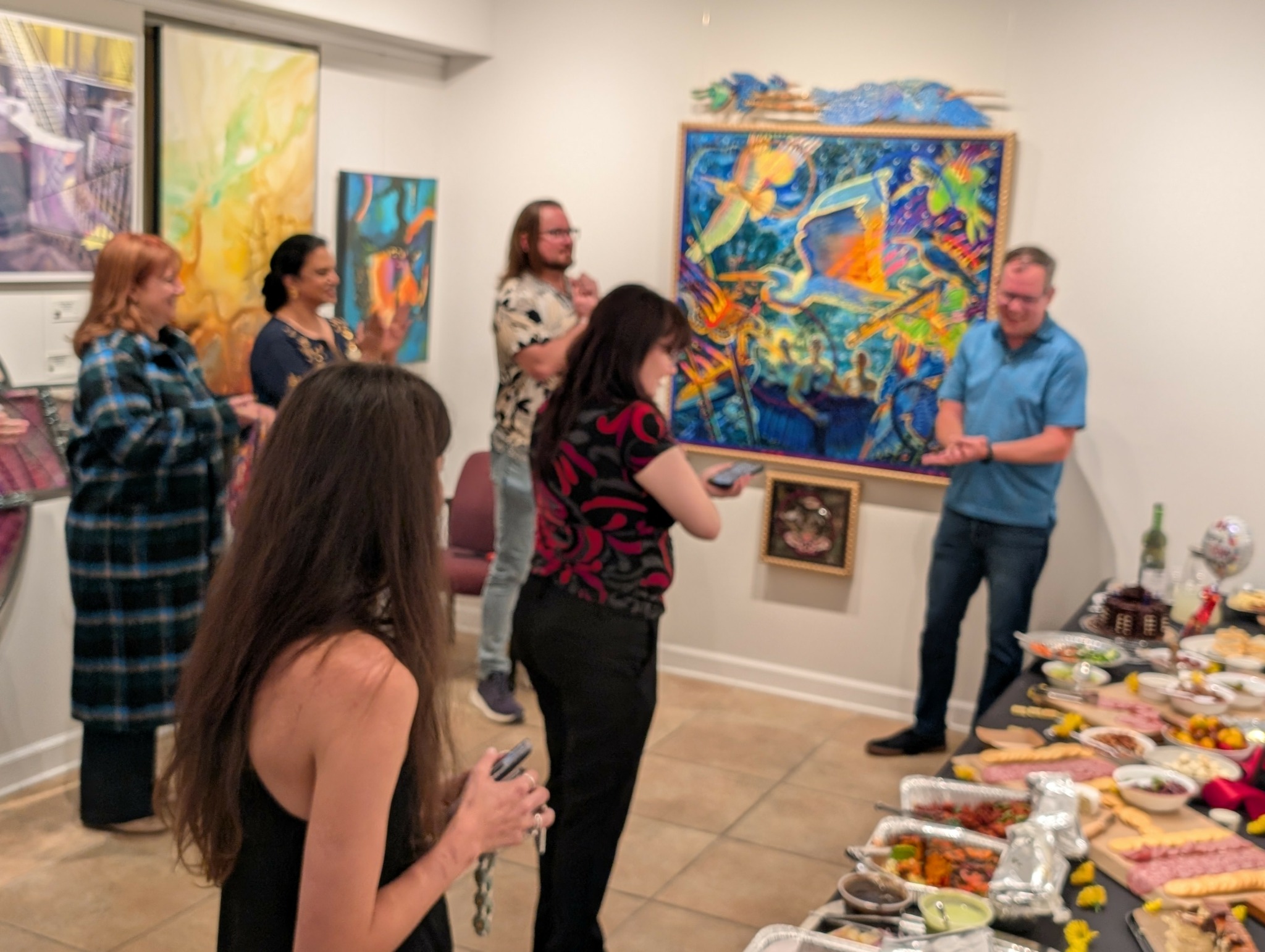 Group of people in an art gallery, some looking at paintings, one person speaking, table with food and drinks.