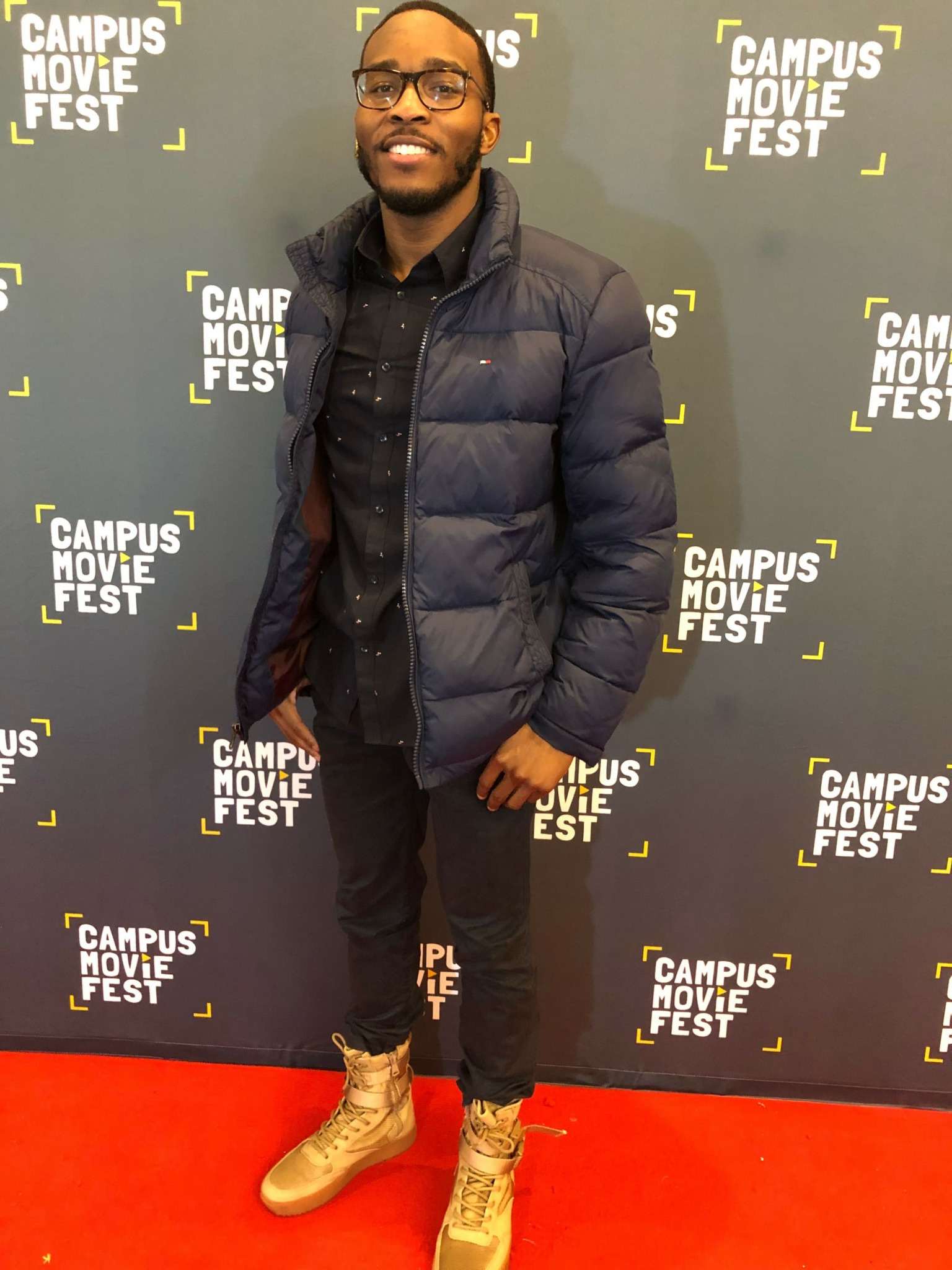 Man in glasses, black jacket, and jeans standing on red carpet with black backdrop, smiling.