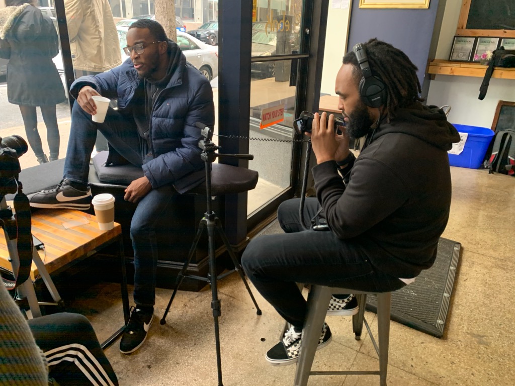 Two men sit indoors near large windows, one with a coffee cup, the other with headphones, engaged in conversation.