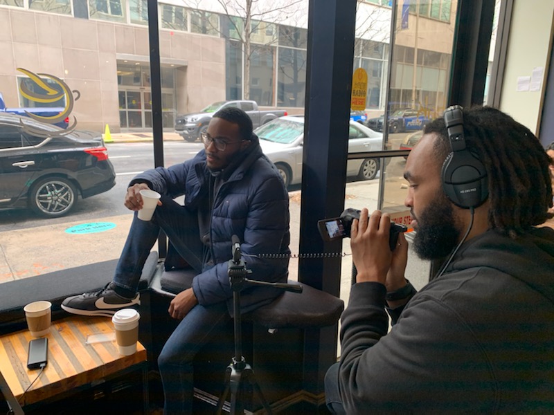 Two men sit indoors near large windows, one with headphones speaking into a microphone, the other holding a cup.