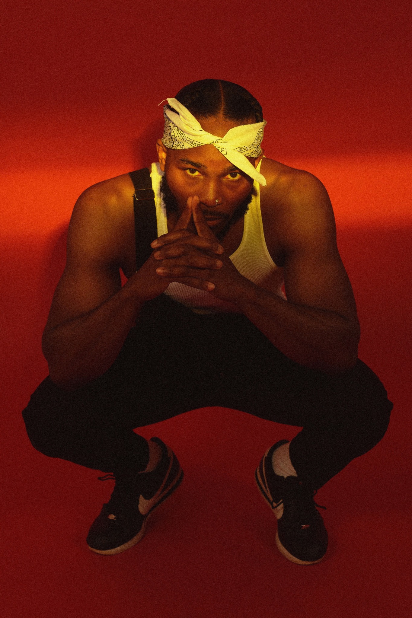 Person crouching with hands clasped, wearing a yellow headband and sleeveless top, against a red background.