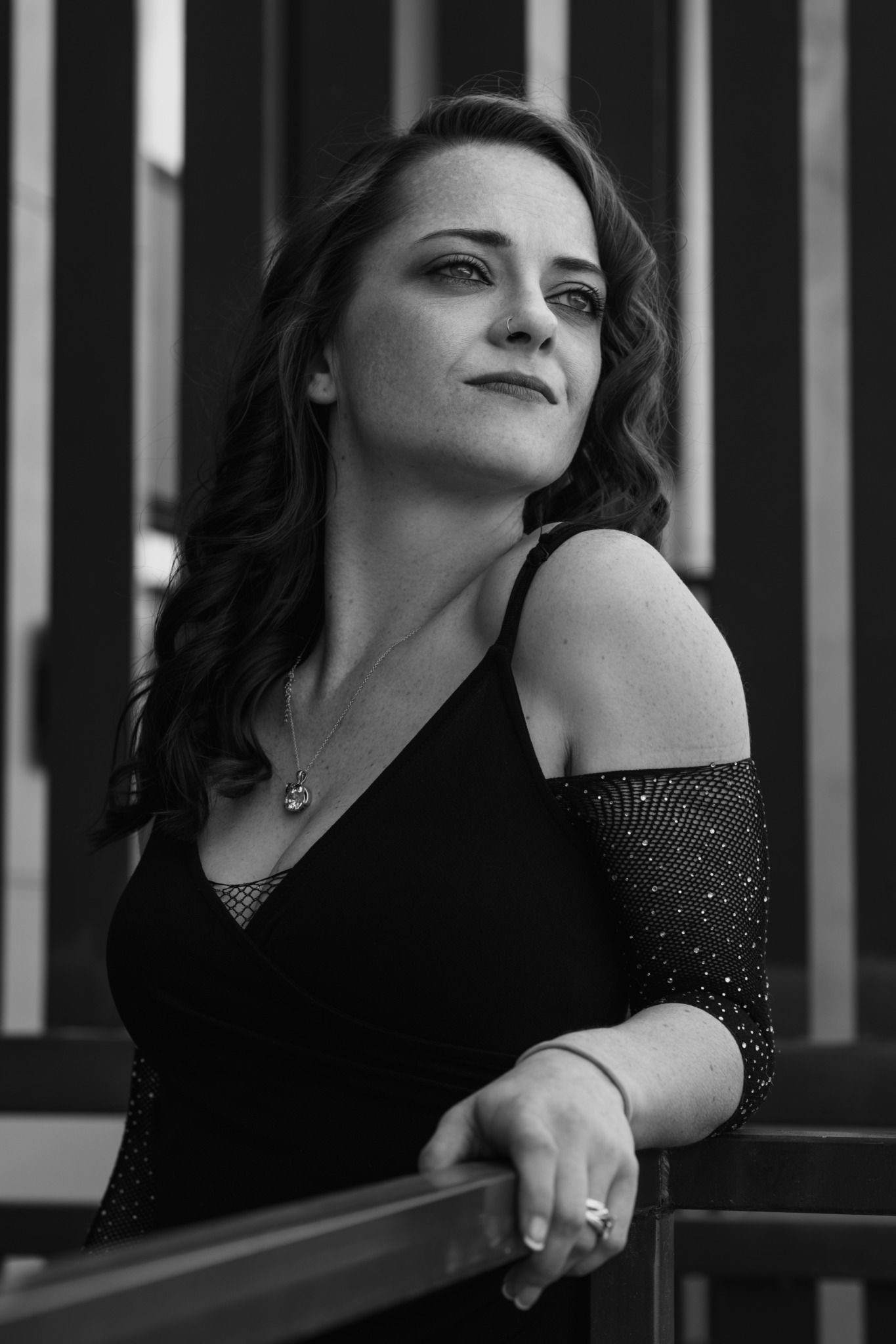 Woman with wavy hair in a black dress, standing outdoors near vertical bars, looking to the side.