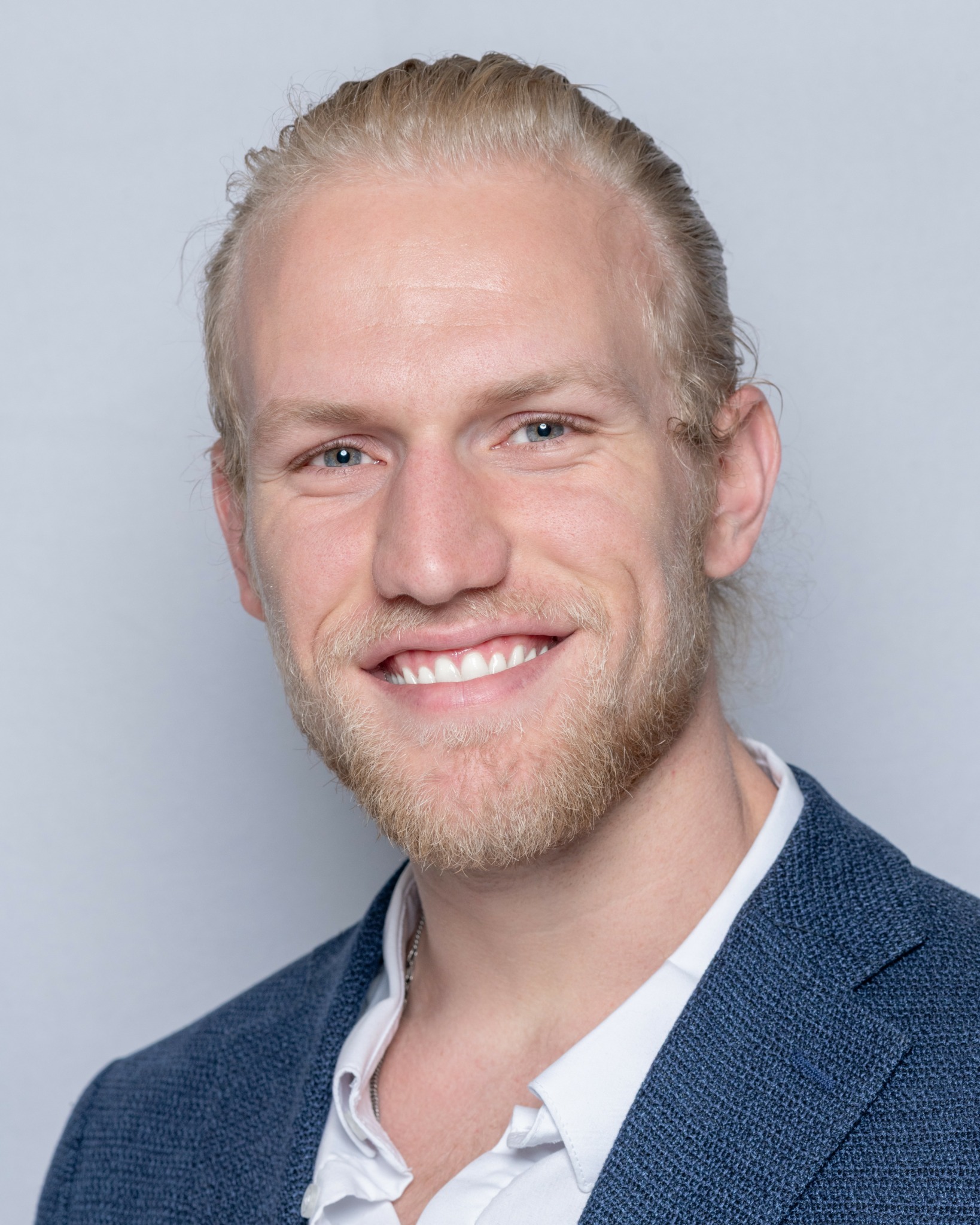 Smiling man with blond hair, beard, wearing a blue blazer and white shirt, against light background.