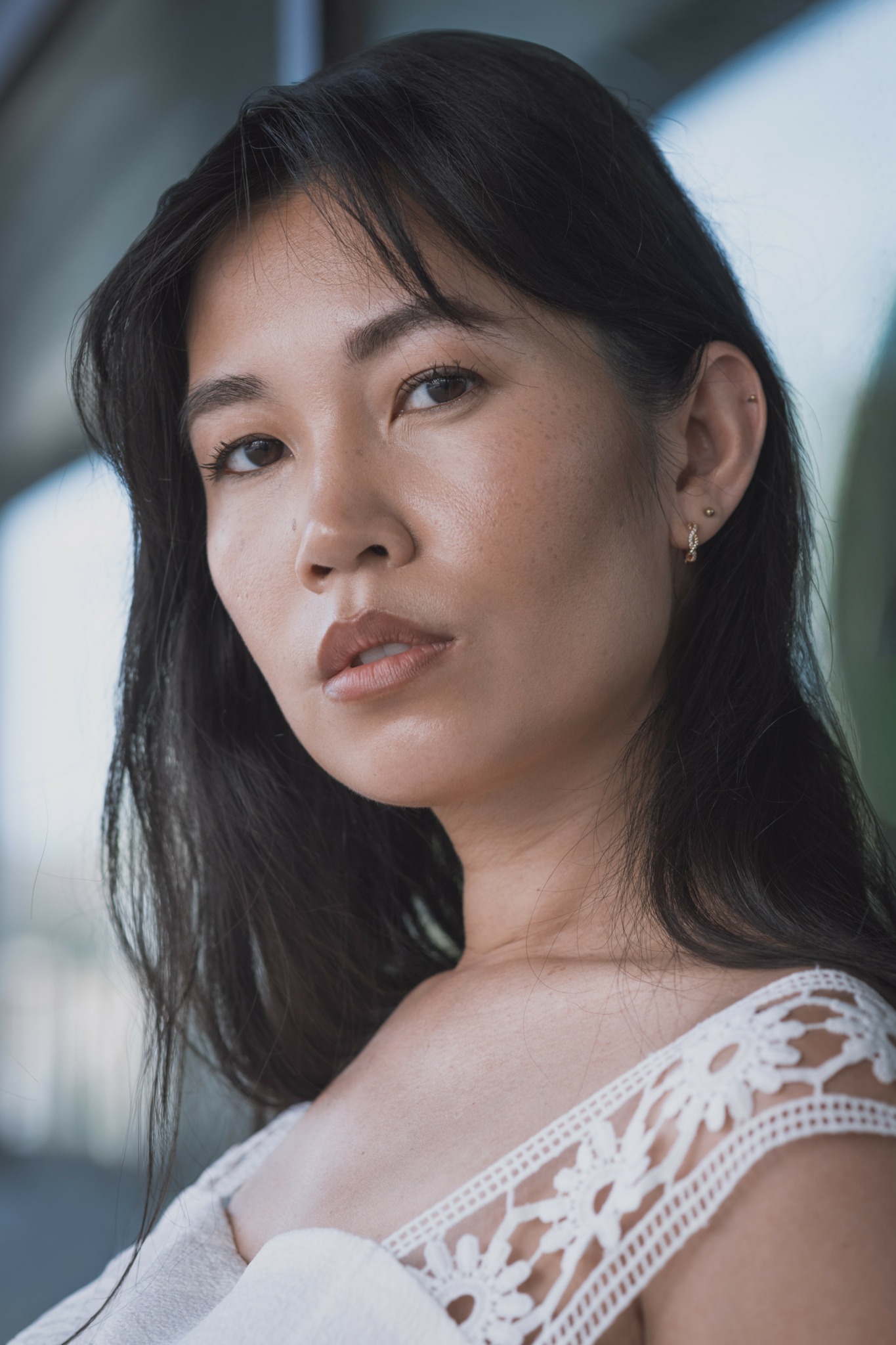 Close-up of a woman with dark hair and earrings, wearing a white lace top, looking at the camera.
