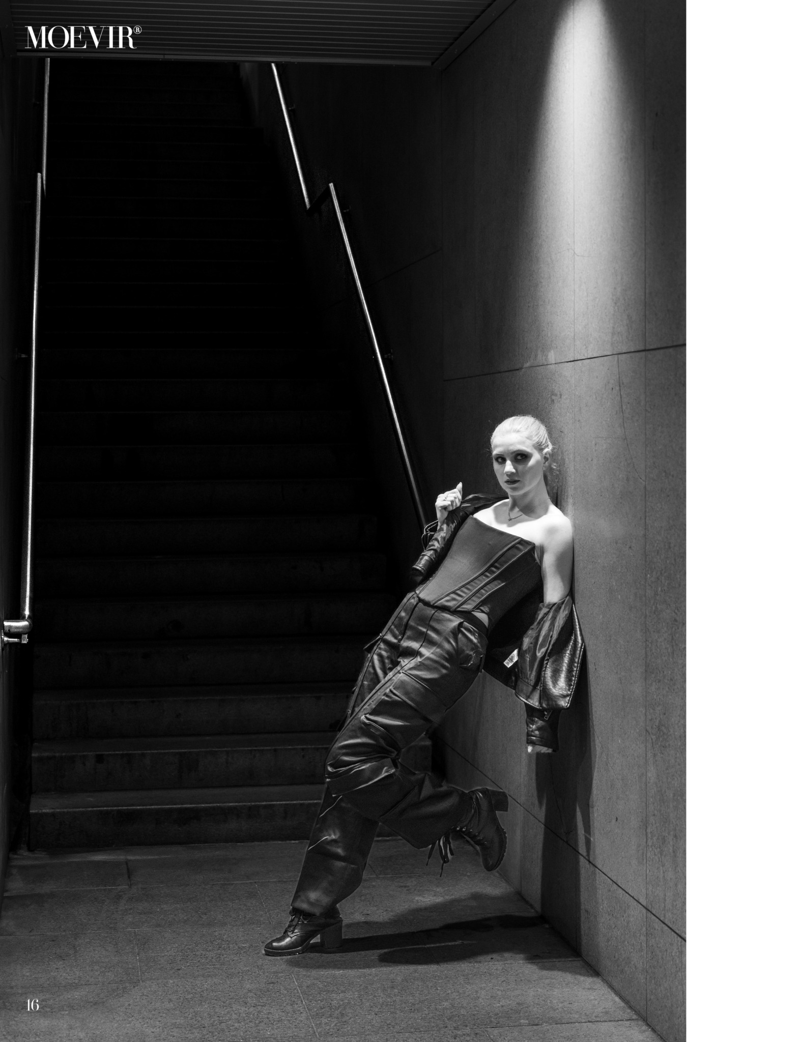 Person leaning against wall near staircase, wearing dark clothing, in a dimly lit space.