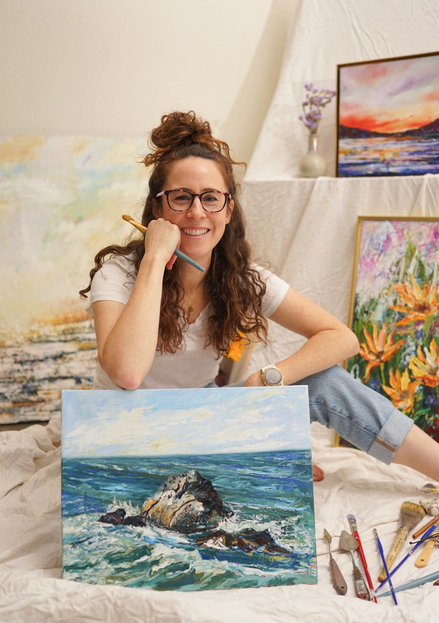Woman with glasses and curly hair sitting among colorful paintings, smiling, with a painting of a rocky ocean scene in front.