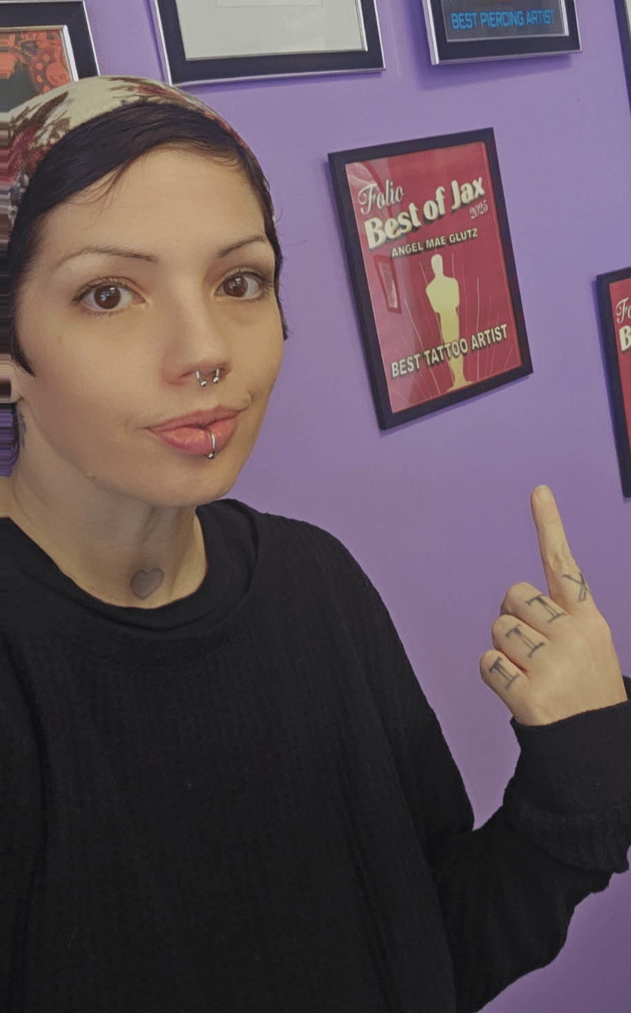 Person with short dark hair and piercings pointing at a framed poster on purple wall.