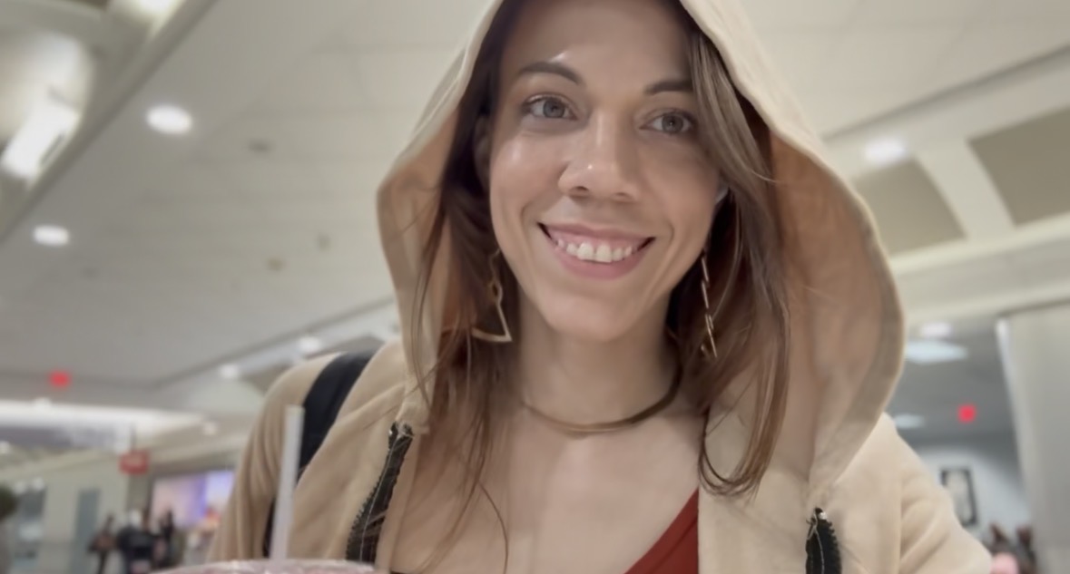 Woman smiling in an indoor public space, wearing a beige hoodie and red top.