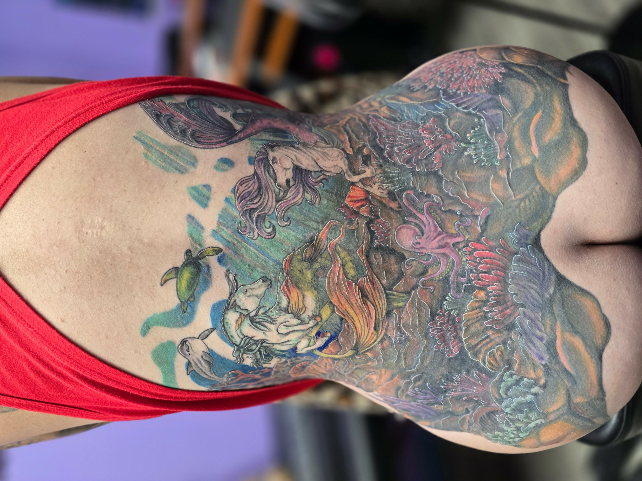 Back of person with colorful tattoos covering shoulders and upper back, wearing a red top.