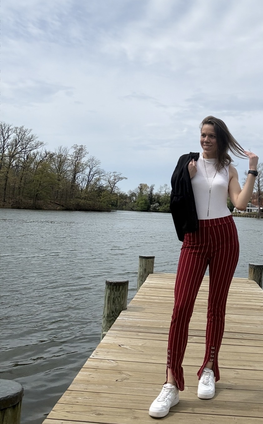 Woman standing on a wooden dock by a body of water, wearing striped pants and white sneakers, with trees and sky in background.