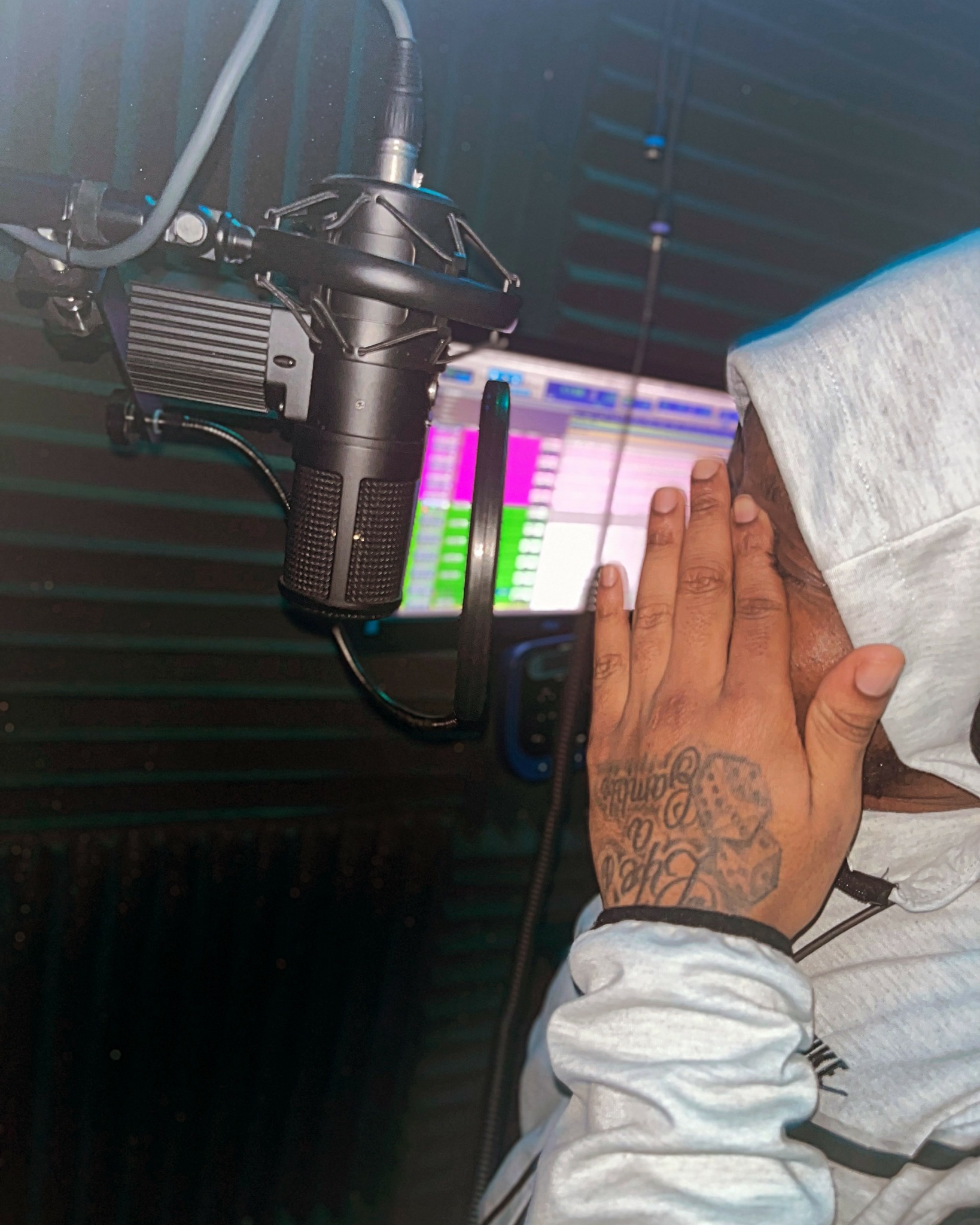 Person's hand with tattoo on wrist near a microphone and a computer screen with colorful audio editing software.