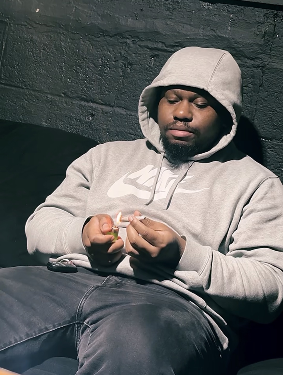 Young man in gray hoodie sitting against a dark brick wall, looking at his hands.