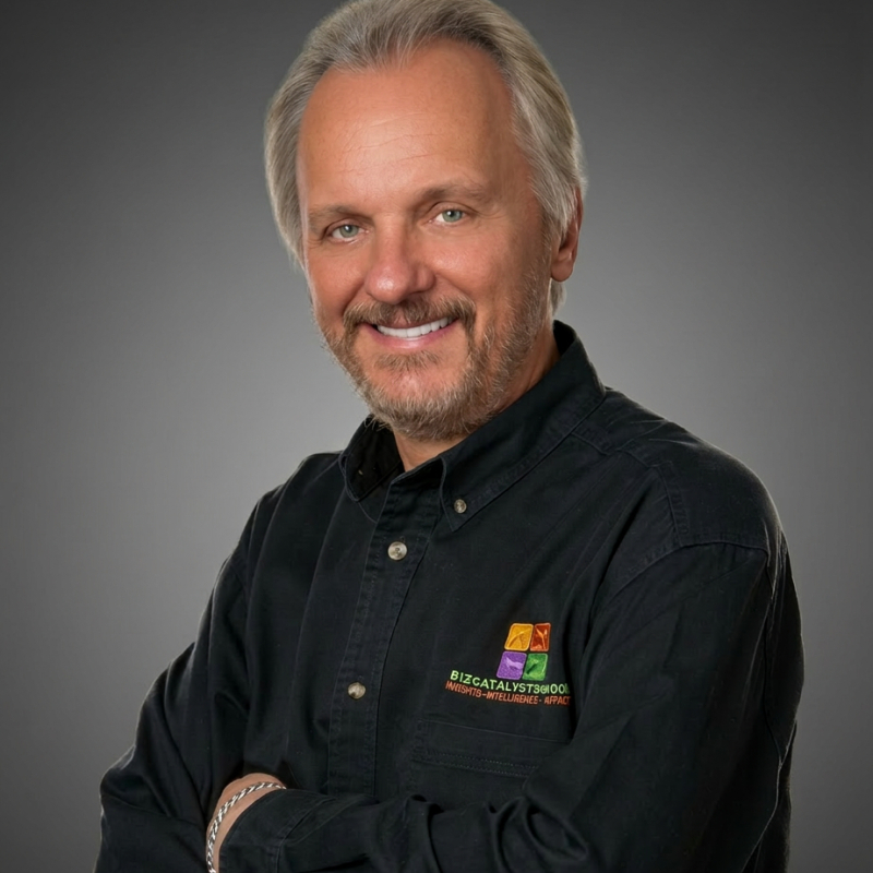 Man with gray hair and beard wearing a black shirt with a logo, smiling against a gray background.