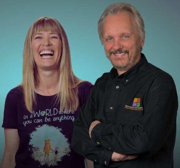 A smiling woman with long blonde hair and a man with gray hair and beard stand together against a blue background.